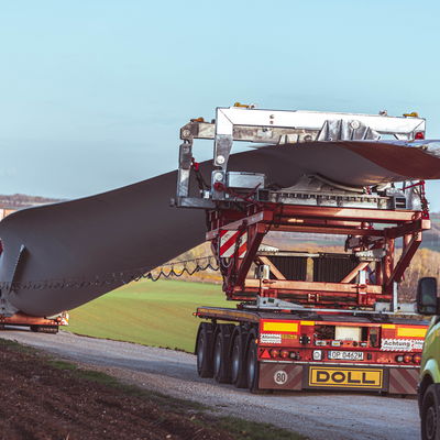 Transport lourd d'une pale d'éolienne sur un camion-remorque spécialisé de DOLL sur une route de campagne.
