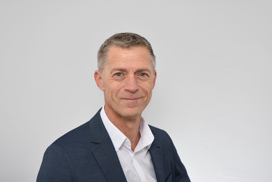 Mann mittleren Alters mit kurzem grauen Haar, im marineblauen Anzug und weißen Hemd, lächelt vor weißem Hintergrund.