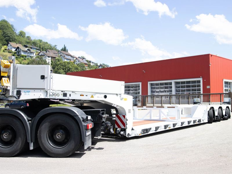 Großer weißer Tieflader an Mercedes-Benz-LKW gekoppelt, geparkt vor einer roten Industriehalle.
