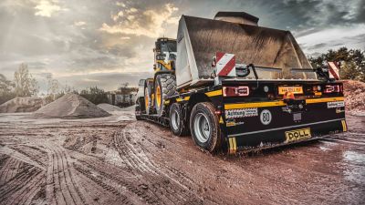 Remorque surbaissée DOLL sur un chantier boueux entre des tas de terre, chargée d'un véhicule de chantier