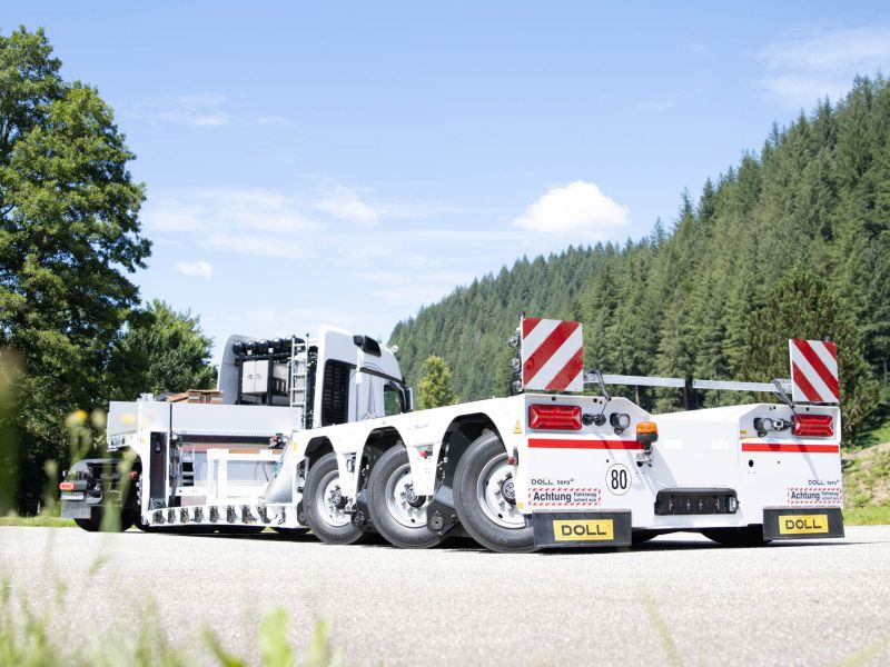 Weißer Tieflader mit Warnmarkierungen, an einen LKW gekoppelt, auf einer Straße im Wald geparkt.