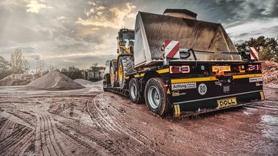 Remorque surbaissée DOLL sur un chantier boueux entre des tas de terre, chargée d'un véhicule de chantier