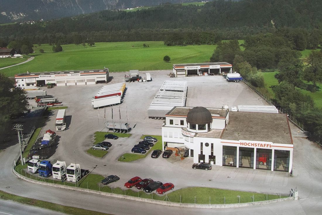 Luftaufnahme eines Nutzfahrzeughändlers mit parkenden LKWs und Anhängern, umgeben von grüner Landschaft und Waldgebiet.