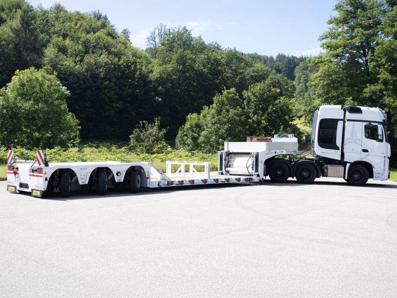 Weißer Tieflader mit Warnmarkierungen, an einen LKW gekoppelt, auf einer Straße im Wald geparkt.